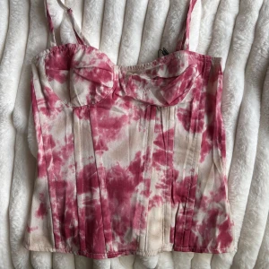 Rosa och beige korsett från PrettyLittleThing - Supersnygg korsett-topp med smala axelband från PrettyLittleThing. Mönstret är batik i rosa och beige toner, med markerade sömmar och bystparti. Toppen har dragkedja bak och är perfekt för att skapa en trendig look. 