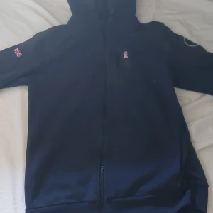 Mörkblå Sail Racing vindjacka med huva - Mörkblå vindjacka från Sail Racing med huva, hel dragkedja och snygga Union Jack-detaljer på bröstet och ärmen. Jackan har en diskret bröstficka med dragkedja och en rund 77-logga på ena ärmen. Perfekt för dig som gillar sportig och stilren look. 160-175cm. Knappt använd.