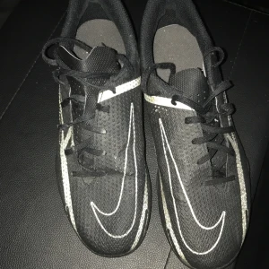 Svarta Nike fotbollsskor med snörning - Svarta fotbollsskor från Nike med vit swoosh längs sidan och snörning framtill. Ovandelen är i syntetmaterial med meshstruktur för bra ventilation. Skorna har en sportig look och passar perfekt för dig som älskar fotboll.
