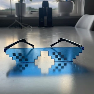 Pixelade blå solglasögon - Pixelinspirerade solglasögon med blåtonade linser och svarta skalmar. Glasögonen har en kantig, stegad form som påminner om 8-bitars grafik och svarta rutmönster på linserna. Perfekta för dig som gillar gaming eller vill sticka ut med en unik stil.