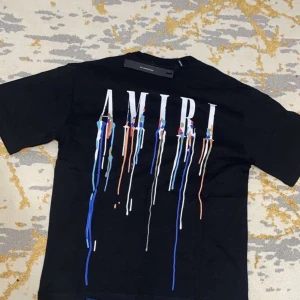 Svart Amiri t-shirt med färgstänk - Cool svart t-shirt från Amiri med stort vitt tryck på ryggen och färgstänk i blått, vitt och orange som rinner ner. Klassisk passform och korta ärmar, perfekt för dig som gillar streetwear med edge.