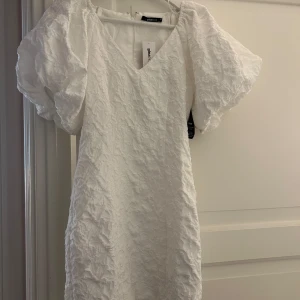 Vit kort klänning från Gina Tricot - Superfin vit kort klänning från Gina Tricot med puffiga korta ärmar och v-ringning. Klänningen har ett strukturerat tyg med diskret mönster och dragkedja bak. Perfekt för dig som vill sticka ut med en trendig och elegant look. Prislapp kvar 