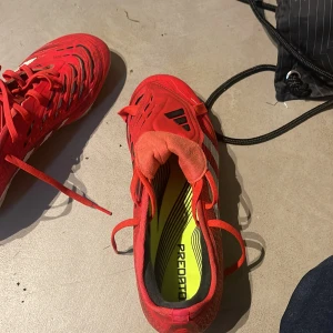 Adidas Predator röda fotbollsskor - Säljer ett par Adidas Predator fotbollsskor i rött och vitt med svarta detaljer. Skorna har snörning, mönstrad ovandel och tydliga dobbar för bra grepp på planen. Snygg design med klassiska Adidas-ränder och Predator-tryck under sulan.