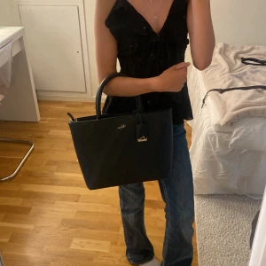 Svart handväska från Rizzo i skinn - Säljer en stilren svart handväska från Rizzo i äkta skinn med guldfärgade detaljer och dragkedja. Väskan har två rejäla handtag och flera fack inuti för att hålla ordning på dina grejer. 