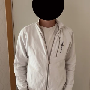 Vit zipjacka från Sail Racing - Vit jacka från Sail Racing med hel dragkedja och ståkrage. Jackan har en bröstficka med dragkedja och logga på vänster sida. Tillverkad i mjukt bomullsmaterial och har långa ärmar med muddar. Perfekt för en clean och sportig stil.