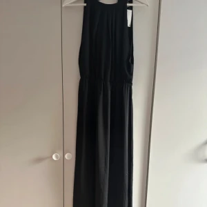 Svart långklänning med spetsdetaljer - Snygg svart långklänning med öppen rygg och vackra spetsdetaljer längs ryggen. Klänningen har ärmlös design och markerad midja, vilket ger en elegant och stilren look