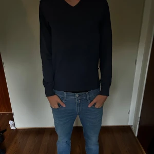 Svart stickad tröja från Hugo Boss - Svart stickad tröja från Hugo Boss med v-ringad hals och diskret logga på bröstet. Tillverkad av fin italiensk ull, perfekt för lager-på-lager och snygg till både jeans och chinos. Klassisk passform och långa ärmar.