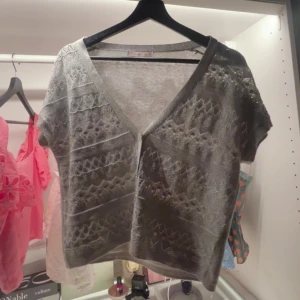 Grå stickad croppad blus med knappar - Superfin grå stickad blus med korta ärmar och v-ringning. Den är croppad och har snygga knappar framtill. Mönstret är genombrutet med coola geometriska detaljer som ger en trendig look. Perfekt att styla med jeans eller kjol för en chill vibe.