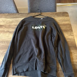 Svart Levi's sweatshirt med camo-logga - Svart sweatshirt från Levi's i storlek M. Tröjan har en klassisk passform med rund hals och långa ärmar. På bröstet finns en camo-inspirerad Levi's-logga i vitt och grönt. Perfekt för en avslappnad och cool stil.