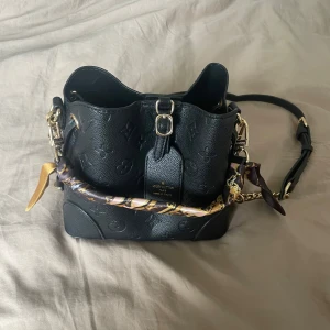 Svart Louis Vuitton axelväska i skinn - Säljer en svart axelväska från Louis Vuitton i präglat skinn med klassiskt monogrammönster. Väskan har guldfärgade metalldetaljer, justerbar axelrem och en snygg scarf-detalj på handtaget. Invändigt finns dragkedjefack för småsaker.