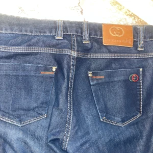 Gucci blå jeans med broderi - Snygga blå jeans från Gucci med klassisk femficksdesign, kontrastsömmar och broderad logga i rött och grönt på bakfickan. Jeansen har läderpatch bak i midjan och detaljer i metall. Perfekta för dig som vill sticka ut med exklusiv streetstyle.