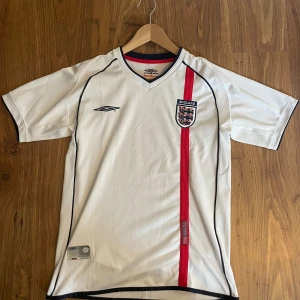 England fotbollströja Umbro vit - Säljer en klassisk vit England fotbollströja från Umbro med röd vertikal rand och broderat landslagsemblem på bröstet. Tröjan har svarta detaljer, V-ringning och är tillverkad i ett lätt, ventilerande material. Perfekt för match eller träning. Tröjan har lite slitage vilket syns på sista bilden .