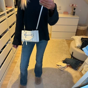 Vit handväska - Snygg vit minihandväska i skinnimitation med både handtag och justerbar axelrem. Väskan har silverfärgade metalldetaljer, en dragkedja framtill och dekorativa knytband. Perfekt storlek för det viktigaste och ger en clean look till outfiten.