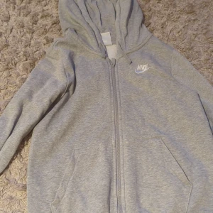 Grå hoodie från Nike med dragkedja - Snygg grå hoodie från Nike med vit logga på bröstet. Tröjan har huva, långa ärmar, två fickor framtill och hel dragkedja. Perfekt för en chill och sportig look. Tillverkad i mjuk bomullsblandning för extra komfort.