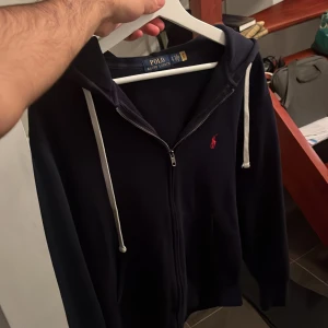 Marinblå hoodie från Polo Ralph Lauren - Säljer en marinblå hoodie från Polo Ralph Lauren med dragkedja och vit snörning i huvan. Mjuk bomullskänsla, perfekt för chill dagar. Kan vara lite dammig, behöver endast avdammas med en klister rulle! Samt lite nopprig men inget som förstör användningen🤩 Nypris 2000 ⬆️ Mvh ItsForfits🌟