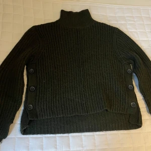 Mörkgrön stickad polotröja med knappar - Mysig mörkgrön stickad polotröja med hög krage och snygga knappar längs båda sidorna. Tröjan har lång ärm och en chunky ribbad struktur som ger en cool och avslappnad vibe. Perfekt för kyliga dagar när du vill vara både varm och stilren.