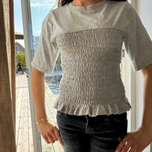 Grå smockad topp med volangkant - Grå topp med smockad midja och volangkant nertill. T-shirtrund hals, halvkort ärm och stretchigt material som sitter snyggt och kan passa upp till storlek M/L.Perfekt för dig som gillar en enkel men ändå söt stil. Använd fåtal gånger. Säljer för den ej kommer tills användning. 