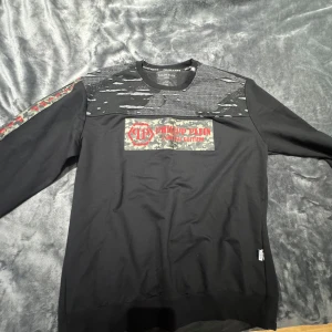 Svart Philipp Plein tröja med camo-detaljer - Svart långärmad tröja från Philipp Plein med coola camo-detaljer och röd logga på bröstet. Unik design med slitna partier upptill och camo-band med röd text längs ärmarna. Perfekt för dig som vill sticka ut med streetstyle-vibe.