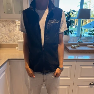 fleeceväst från Peak Performance - Svart fleeceväst från Peak Performance med hög krage och dragkedja framtill. Västen har en bröstficka i blankare material med logga och en clean, sportig look. Perfekt lager-på-lager-plagg för kyliga dagar och sommar kvällar