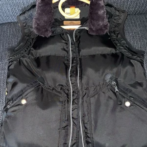 Parajumpers kobuk väst  - Svart dunväst från Parajumpers med fluffig krage, dragkedja framtill och flera fickor med dragkedja. Västen har quiltad design och detaljer i gult på insidan. Perfekt för lager-på-lager under kyliga dagar.