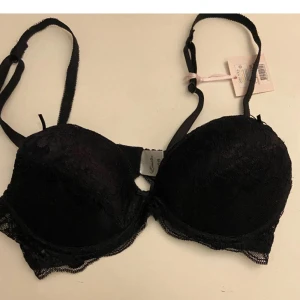 Svart spetsig bh från Emilia Lingerie - Snygg svart bh från Emilia Lingerie med spetsdetaljer och justerbara axelband. Kuporna är lätt vadderade och har en elegant spetskant runtom. Perfekt för dig som vill ha en feminin och stilren look under kläderna.