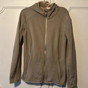 Beige fleecejacka med huva - Mysig beige fleecejacka med huva och hel dragkedja framtill. Jackan har diskreta ribbade paneler på sidorna och två fickor med dragkedja. Perfekt för kyliga dagar och enkel att matcha med det mesta.