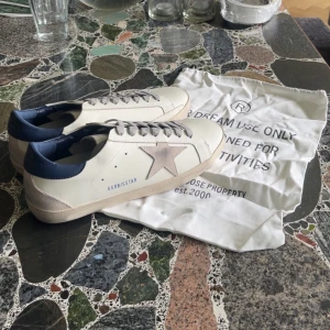 Golden Goose vita sneakers med blå detalj Storlek 42 - Golden Goose sneakers i vitt skinn med grå mockadetaljer och en ikonisk stjärna på sidan. Blå hälkappa i skinn, grå snörning och beige sula. Snygg och stilren modell som passar perfekt till jeans eller kjol. Kommer utan Skokartong