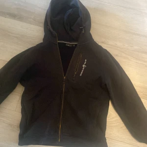 Svart Sail Racing hoodie  med luva  - Svart jacka från Sail Racing med huva och dragkedja framtill. Jackan har en bröstficka med dragkedja och broderad logga på både bröstet och huvan. Perfekt för dig som gillar stilren och sportig look.
