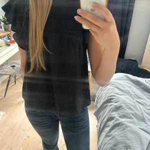 Svart blus med volang från Vero Moda - Supersöt svart blus från Vero Moda i luftig bomull med små broderade prickar. Blusen har volangärmar, hög hals med liten volangkant och fina spetsdetaljer framtill. Perfekt att styla med jeans eller kjol för en trendig look.