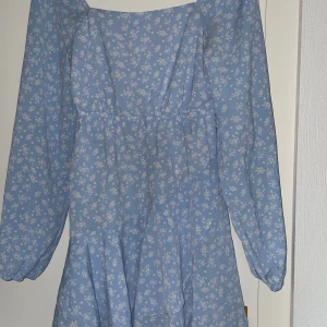 Blå blommig kortklänning med volang - Supersöt ljusblå kortklänning med vita små blommor, långa ärmar och volangdetalj nertill. Klänningen har fyrkantig ringning och smockad rygg för extra bekväm passform. 