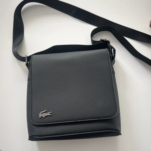 Lacoste axelremsväska - Säljer denna feta axelväska då jag inte använder den. Den är i nyskick utan tecken på användning. Skriv för frågor/bilder