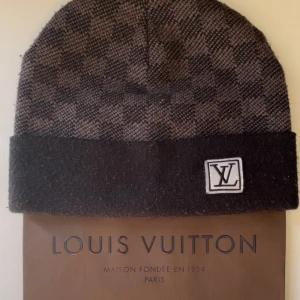 Rutig mössa från Louis V - Snygg rutig mössa i mörkbrunt och svart med klassisk logga framtill. Mössan har uppvikt kant och är tillverkad i mjukt ullmaterial, perfekt för kyliga dagar. En exklusiv accessoar som lyfter din stil.