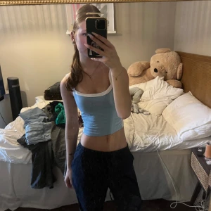 Ljusblå croppad linnetopp med vita kanter - Säljer en ljusblå croppad linnetopp med breda vita kanter och axelband. Toppen har en tight passform och ärmlös design, perfekt för varma dagar. Mjuk och stretchig bomullskänsla som sitter snyggt på kroppen.