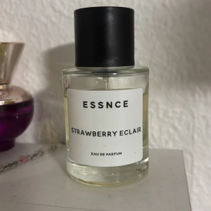 ESSNCE Strawberry Eclair parfym - Parfymen ESSNCE Strawberry Eclair kommer i en stilren, rund glasflaska med svart lock. Doften är inspirerad av jordgubb och bakverk, vilket ger en söt och fruktig känsla. Flaskan är transparent och visar den ljusgula vätskan inuti. Perfekt för dig som gillar unika och söta dofter.