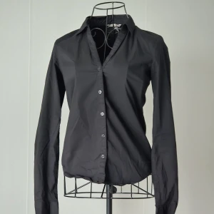 Svart klassisk skjorta med knappar - En stilren svart skjorta med lång ärm och klassisk krage. Skjortan har knäppning framtill med diskreta knappar och en figurnära passform. Materialet känns lätt och smidigt mot kroppen.