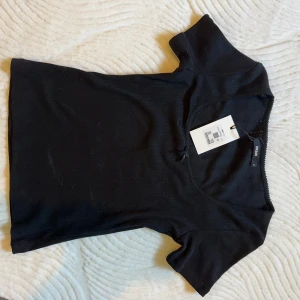 Svart ribbad topp med rosett från Bik Bok - Supersöt svart ribbad topp från Bik Bok med kort ärm och liten rosett framtill. Toppen har v-ringning och diskret spetskant längs halsringningen. Perfekt att styla med jeans eller kjol för en trendig look. Ny pris 199kr. 