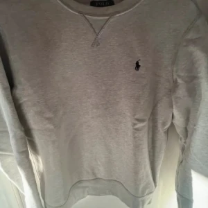 Ralph lauren tröja - Snygg grå sweatshirt från Polo Ralph Lauren med klassisk rund hals och broderad logga på bröstet. Tröjan har långa ärmar och ribbade muddar vid ärmslut och nederkant. Perfekt för en chill och stilren look.