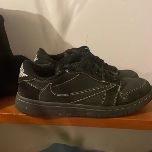 Svarta Nike Air Jordan 1 Low Travis Scott - Svarta Nike Air Jordan 1 Low Travis Scott med vit kontrastsöm, ikonisk omvänd swoosh och broderade detaljer på hälen. Skorna har snörning, rund tå och platt sula. Materialet är skinn och textil, vilket ger en clean och stilren vibe.