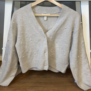 Grå kofta med pärlor från H&M - Superfin ljus beige kofta från H&M med v-ringning och dekorativa pärlor över hela tröjan. Koftan är stickad, har långärmade ärmar och en lite loose passform. Perfekt att slänga över en topp för en extra touch. Tröjan har använts ett par gånger och har ett litet hål i sig som man ser på sista bilden💗