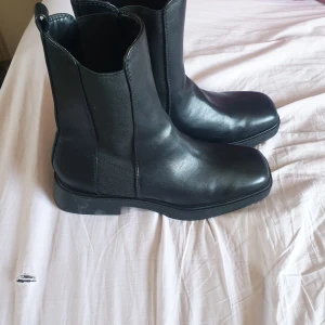Svarta chelsea boots i skinn - Stilrena svarta chelsea boots i skinn med fyrkantig tå och elastiska paneler på sidorna. Klassisk och tidlös modell med platt sula, perfekt till både jeans och kostymbyxor. Snyggt minimalistisk look som funkar året runt. Oanvända då jag köpte fel storlek! Storlek 41