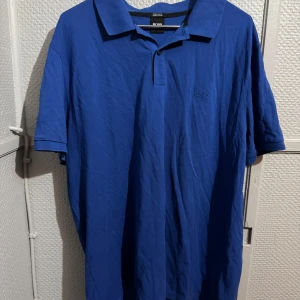 Blå pikétröja från Hugo Boss - Snygg blå pikétröja från Hugo Boss i mjuk Pima-bomull. Klassisk krage, korta ärmar och diskret broderad logga på bröstet. Perfekt för en clean och stilren look. Passar dig som gillar enkel men exklusiv stil.