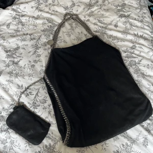 Svartglittrig Stella McCartney Falabella väska - Säljer en svartglittrig axelväska från Stella McCartney med silverfärgade kedjedetaljer och matchande liten plånbok som sitter inuti väskan. Väskan har magnetlås, innerfack med dragkedja och är gjord i syntetmaterial med mockakänsla. Sparsamt använd men syns lite rost på en liten del av kedjan. Skriv för fler bilder. Köpt i Italien för drygt 9 000 kr.