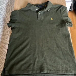 Olivgrön pikétröja från Ralph Lauren - Snygg olivgrön pikétröja från Polo Ralph Lauren i slim fit-modell. Klassisk krage, två knappar och ikoniska broderade loggan i gult på bröstet. Tillverkad i mjuk bomull, perfekt för en clean och stilren look.