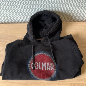 Svart hoodie från Colmar  - Svart hoodie från Colmar med stor rund logga i rött, vitt och blått framtill. Tröjan har huva med snörning och är tillverkad i mjuk bomullsmix. Perfekt för en chill och sportig look. Hoodien e lite trasig vid luvan men annars i riktigt bra skick 
