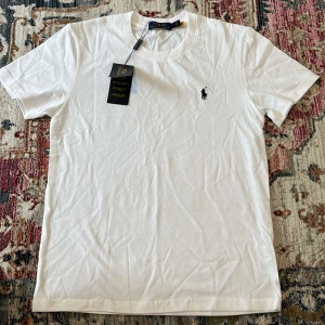 Vit t-shirt från Polo Ralph Lauren - Klassisk vit t-shirt från Polo Ralph Lauren med broderad svart logga på bröstet. T-shirten är i mjuk bomull, har rund hals och korta ärmar. Perfekt basic-plagg med stilren design och snygg passform.