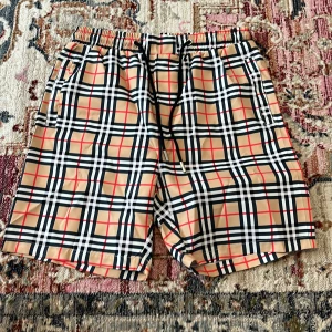 Rutig Burberry shorts med snörning - Snygga shorts från Burberry med klassiskt rutigt mönster i beige, svart, vitt och rött. De har elastisk midja med svart snörning och metalländar, samt sidfickor. Perfekta för sommaren och ger en lyxig vibe till din outfit.