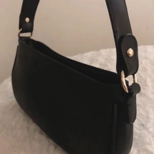 Svart axelväska i skinnimitation - Säljer en stilren svart axelväska med enkel design och bred axelrem. Väskan är tillverkad i skinnimitation och har silvriga metalldetaljer. Perfekt för dig som vill ha en klassisk accessoar till din outfit.