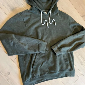 Mörkgrön hoodie från pier one - Säljer en stilren mörkgrön hoodie från Layer One med huva och vita dragsnören. Tröjan har en klassisk känguruficka framtill och ribbade muddar vid ärmslut och nederkant. Perfekt för en avslappnad look. Den är i bra skick överlag inga skador. Skriv vid minsta fundering😁
