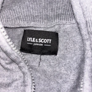 Grå halvzip-tröja från Lyle & Scott Junior - Snygg grå tröja från Lyle & Scott Junior med halv dragkedja och klassisk gul logga på bröstet. Tröjan har lång ärm och ribbade muddar vid hals, ärmslut och nederkant. Perfekt för en stilren och avslappnad look. Storlek 146-152