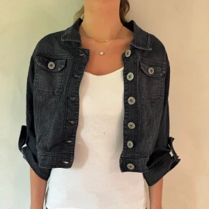 Svart croppad jeansjacka med knappar - Säljer en svart croppad jeansjacka med dekorativa silvriga knappar. Jackan har klassisk krage, långa ärmar och två bröstfickor. Perfekt för dig som vill ha en cool och trendig look.